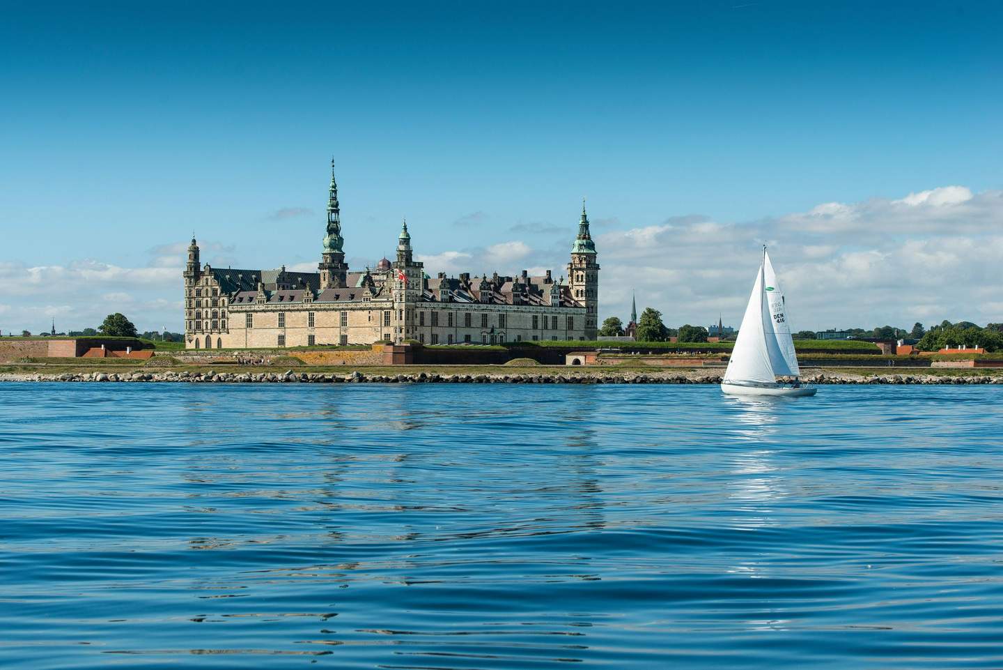 Kronborg Castle A sailboat on calm water with a historic castle featuring multiple spires in the background under a clear blue sky.