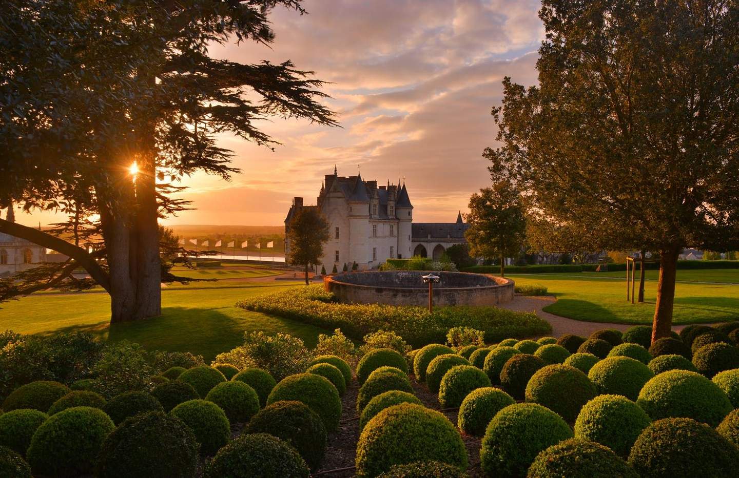 A castle at sunset with neatly trimmed bushes, large trees, and a circular stone structure in the garden.