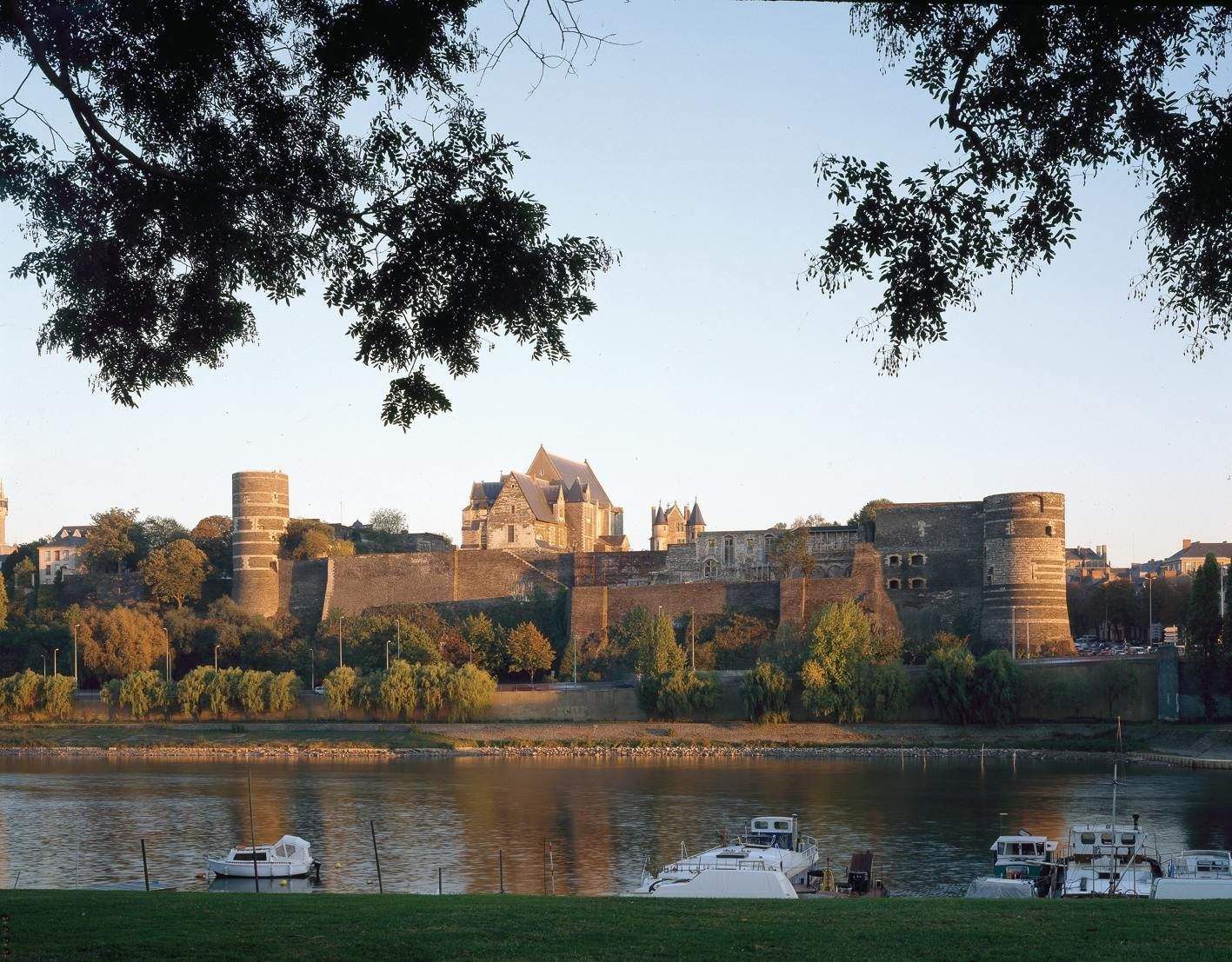 Château d'Angers Ancient stone castle with multiple towers by a river, framed by trees. A few boats are docked along the riverbank.