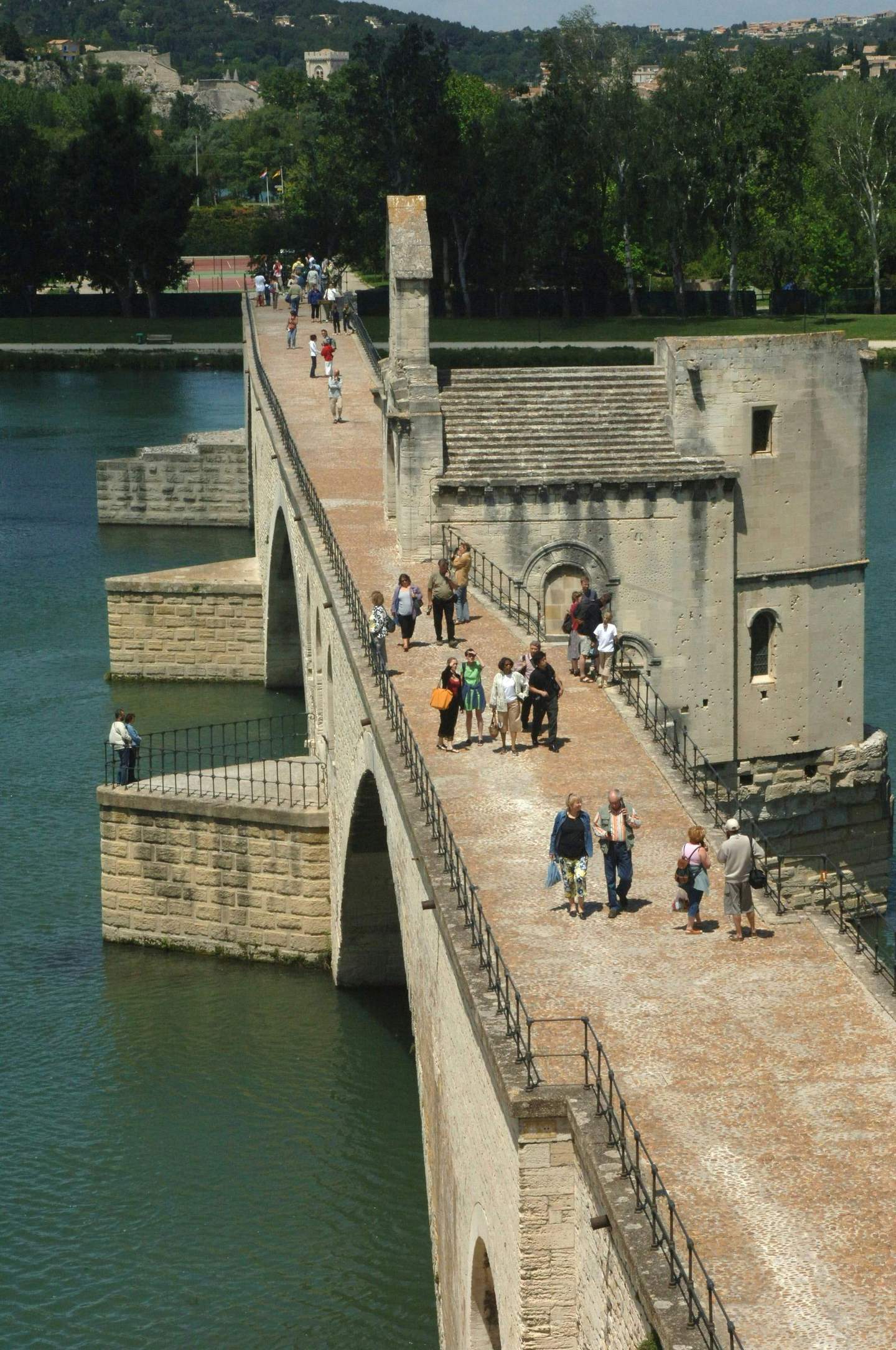 People walking on a historic stone bridge over a river, with trees and buildings visible in the background.
