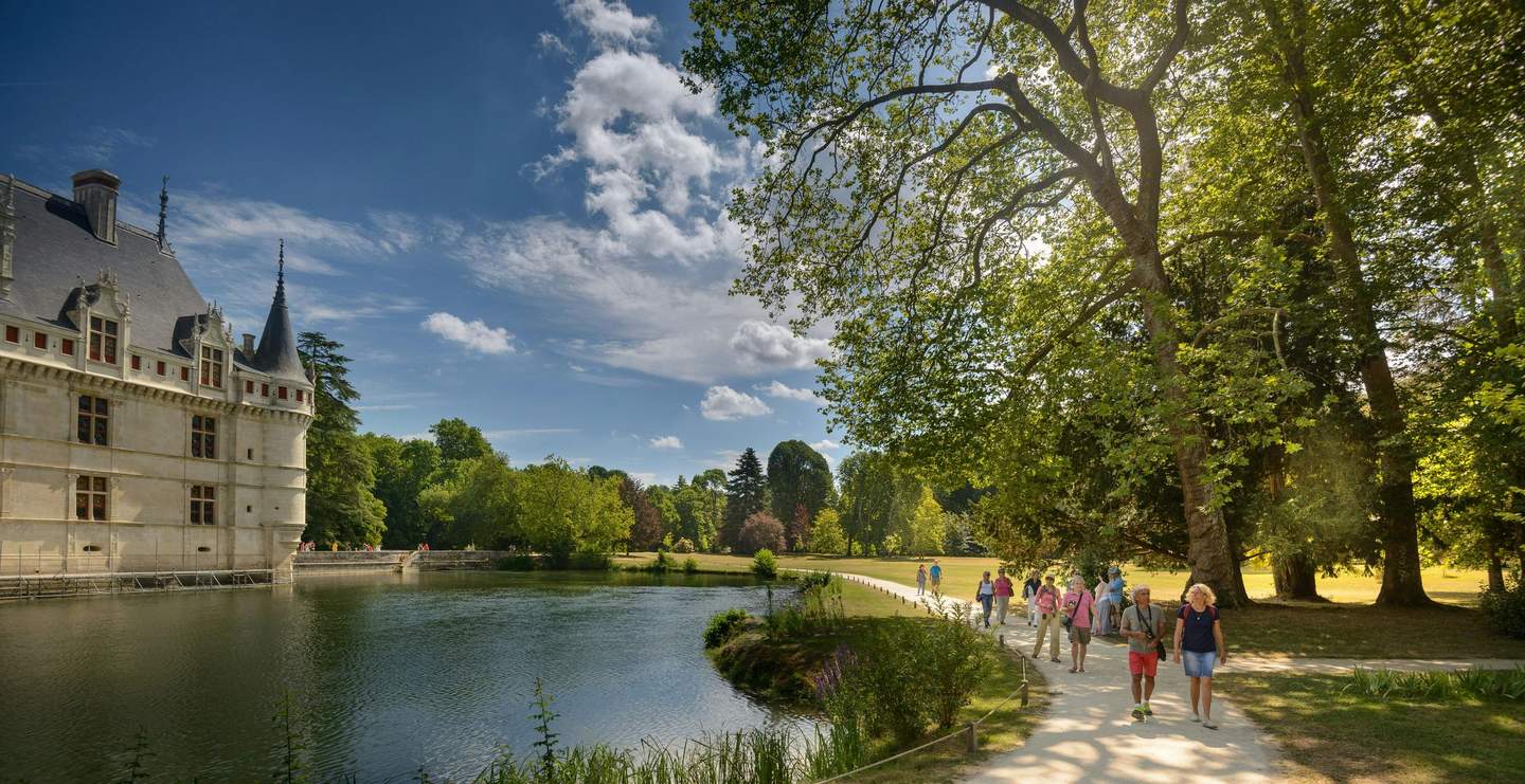 People walking on a path near a tree-lined pond, adjacent to a historic building under a clear, blue sky.