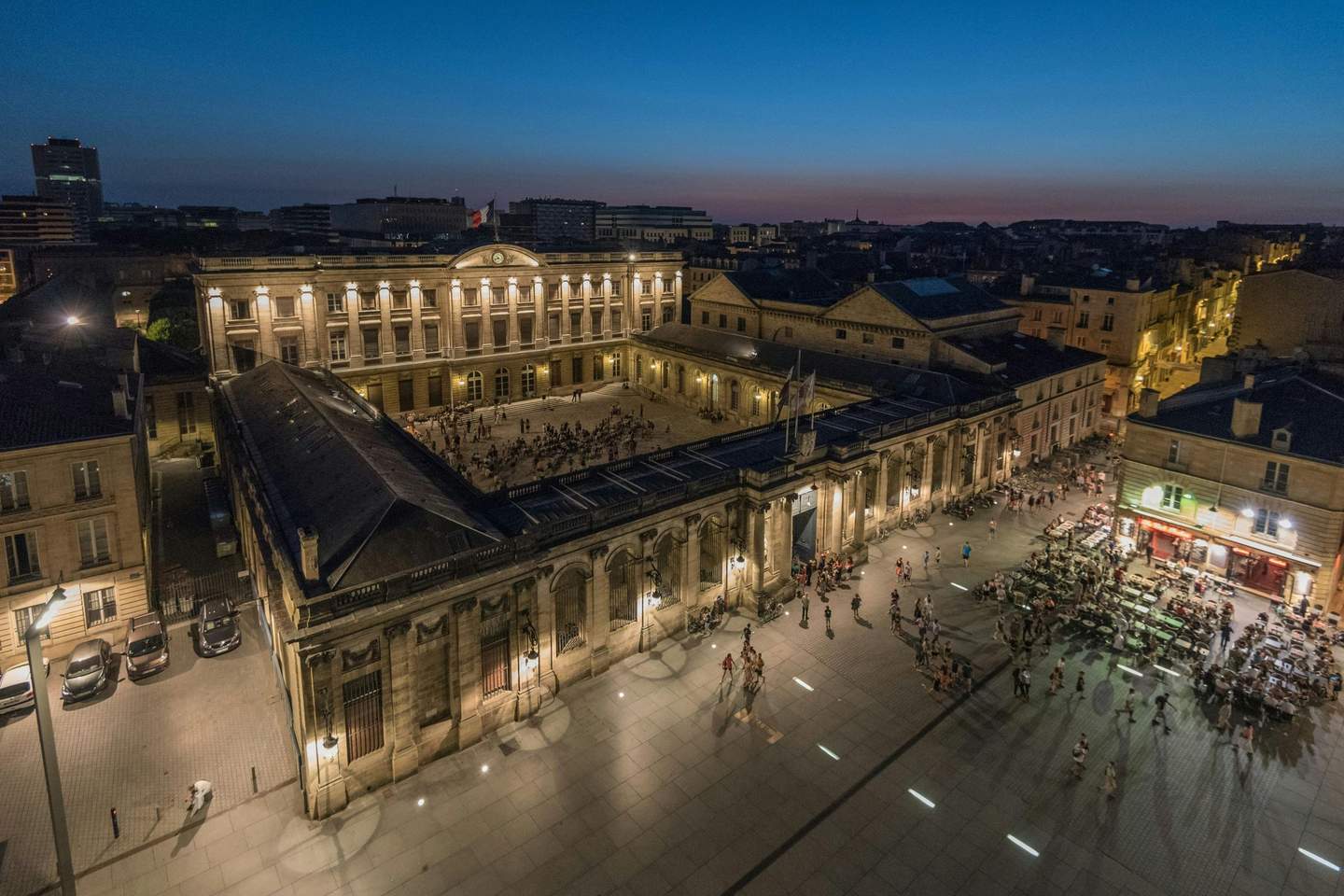 Aerial view of a lit historic building and a courtyard with people at dusk, surrounded by city structures and streets.