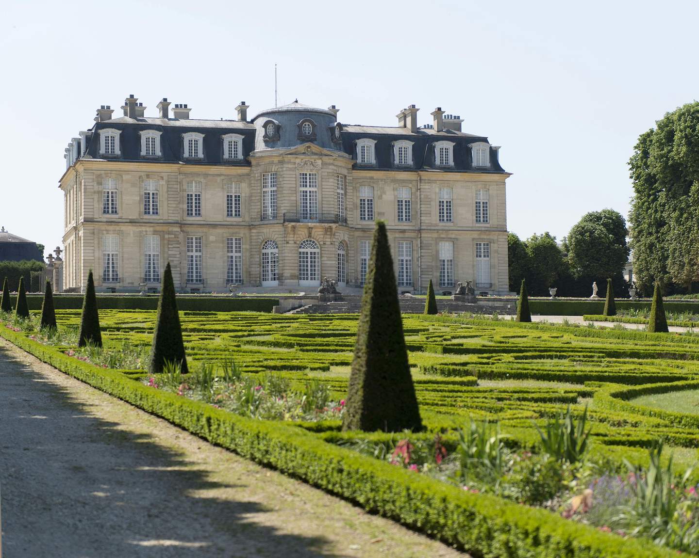 A grand historic building with manicured gardens featuring trimmed hedges and conical shrubs in the foreground.