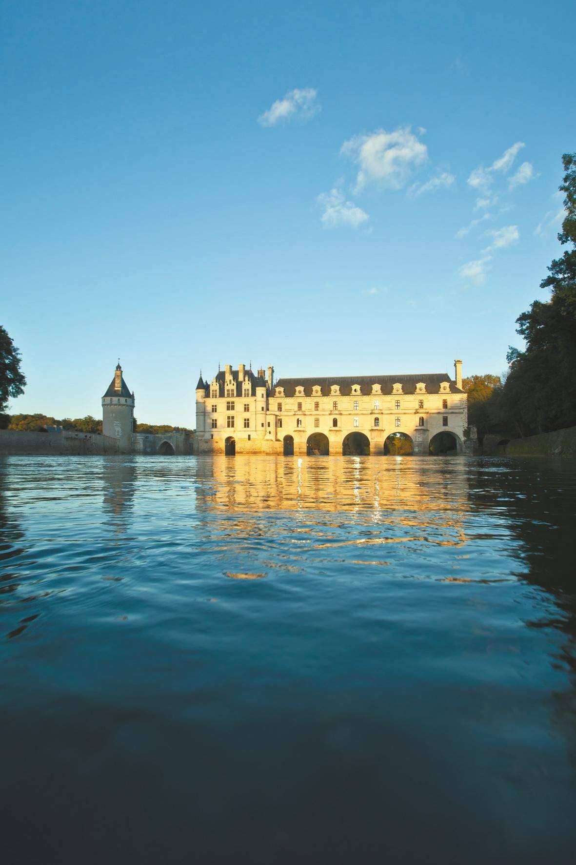 Château de Chenonceau A castle with multiple arches reflected in a calm body of water under a clear blue sky. Trees frame the scene.