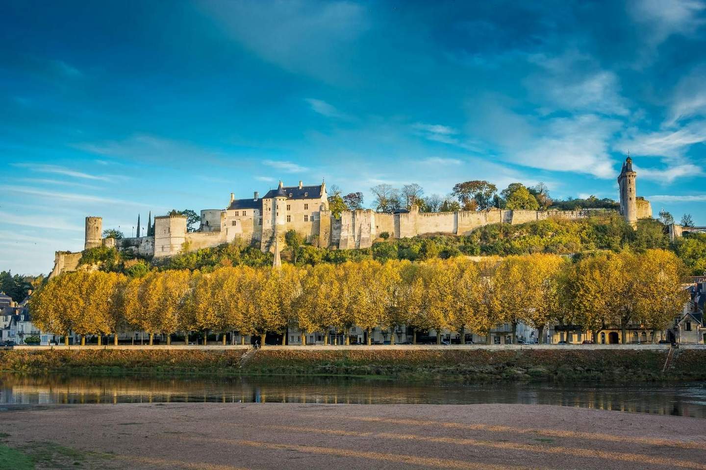 Medieval castle on a hill, surrounded by stone walls, above a line of trees with yellow leaves and a river in the foreground.
