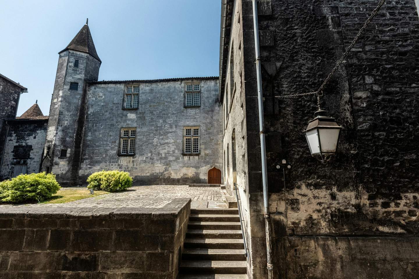 Château Royal de Cognac Stone building with turret and arched door, steps leading up, surrounded by greenery, and an old street lamp on the right.