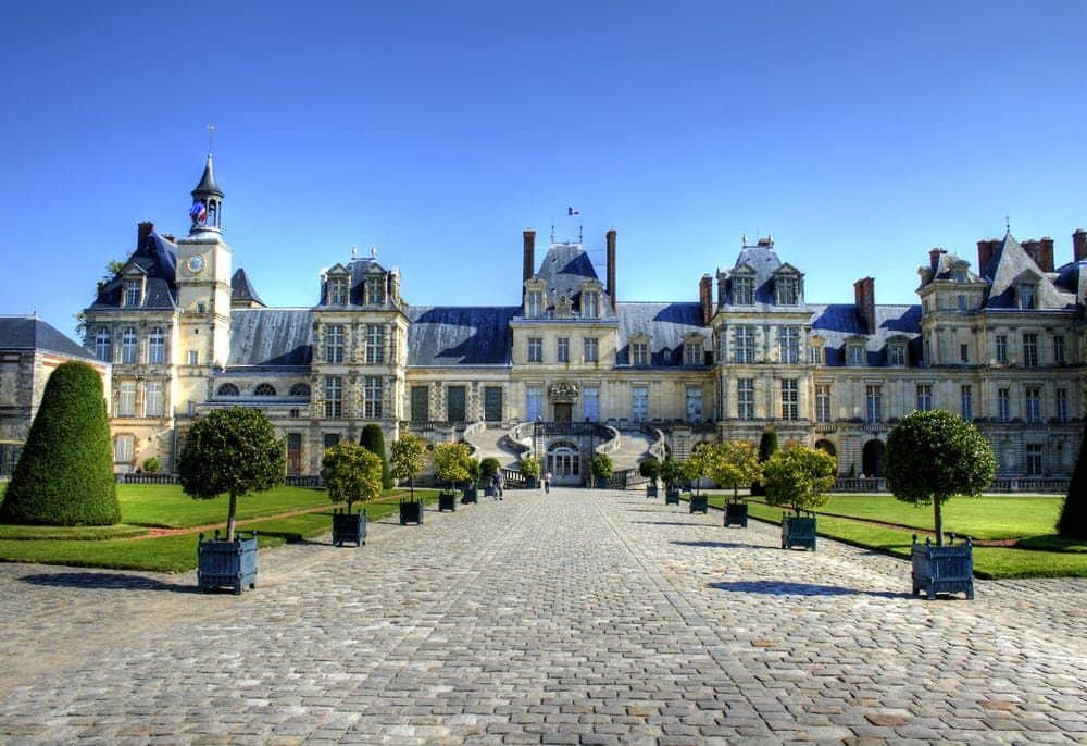 A large historical building with multiple windows and towers, fronted by a cobblestone path and manicured gardens under a clear blue sky.