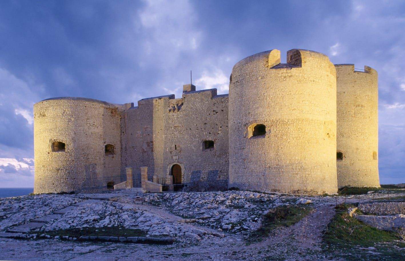 Château d’If A large stone castle with round towers and small windows under a cloudy sky, surrounded by rocky terrain.