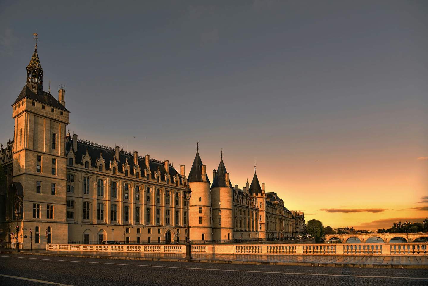 Historic building with tower and turrets along a river at sunset, with a bridge in the background and clear sky.