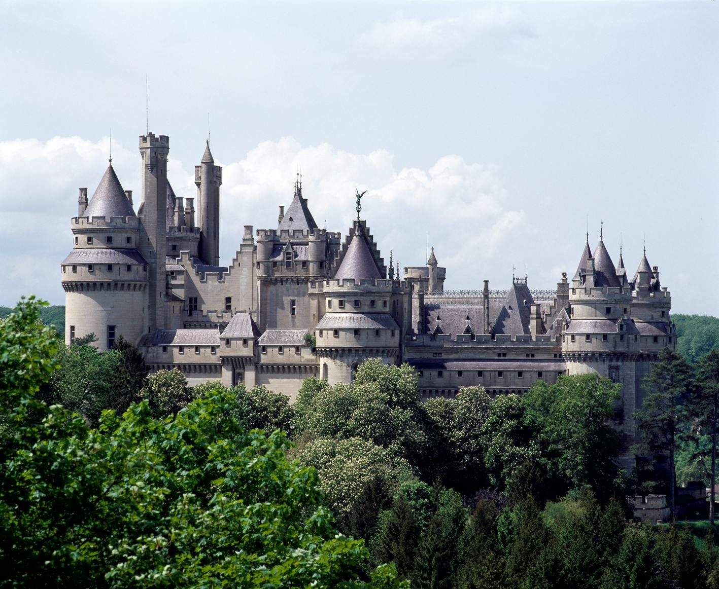 A large, intricate castle with multiple towers and spires surrounded by lush green trees, set against a cloudy sky.