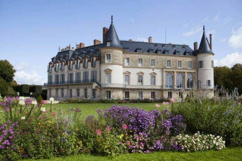A large chateau with gray roofs and round towers is surrounded by colorful flower gardens under a clear blue sky.