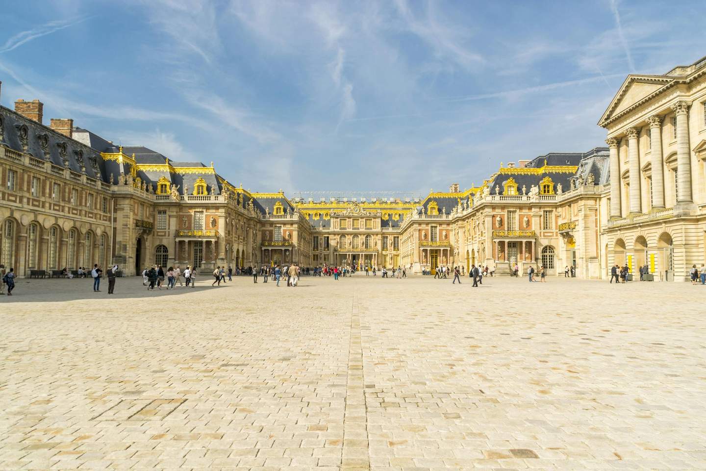 Courtyard of a grand palace with golden decorations on the rooftops. People are walking and taking photos on a sunny day.