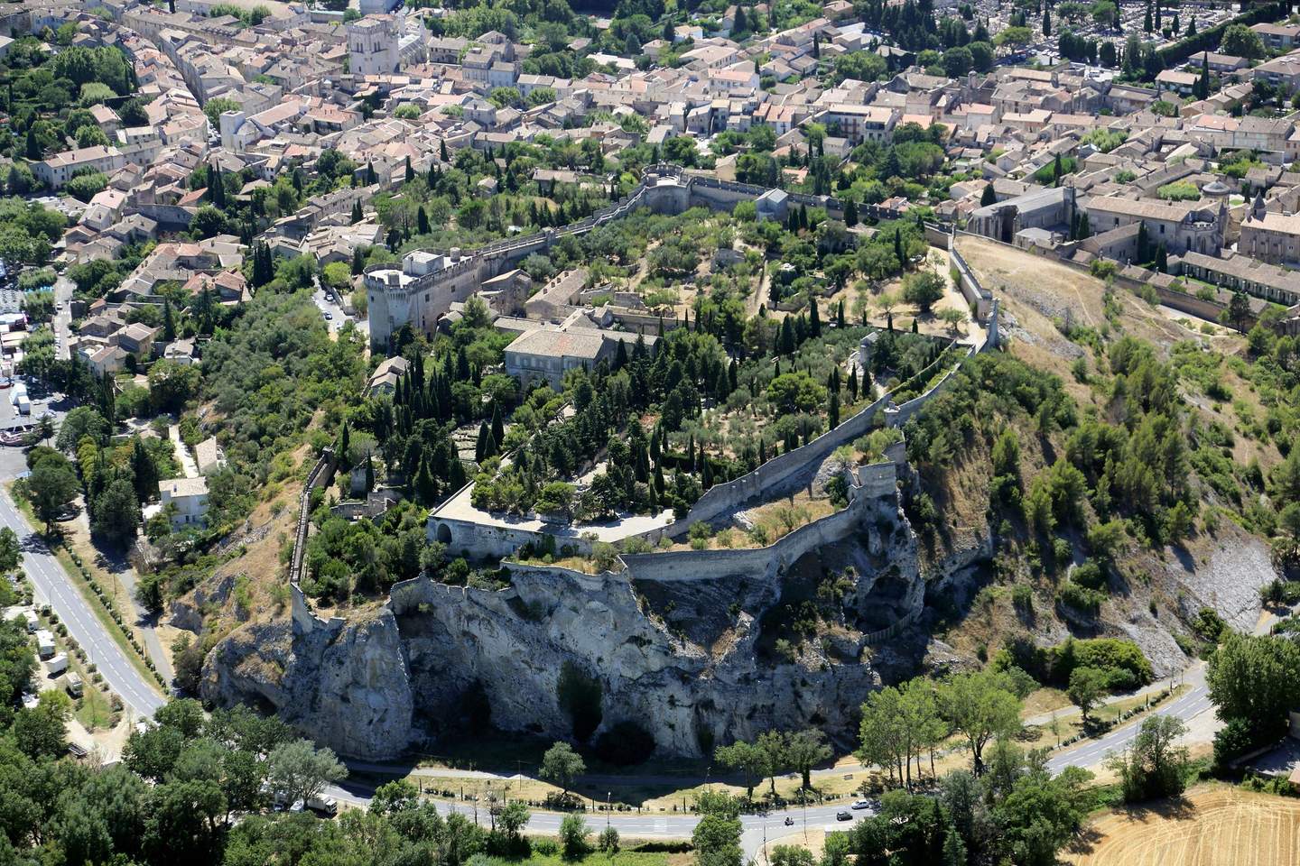 Aerial view of a historical fortified town with stone walls, buildings, and lush greenery surrounded by roads and hills.