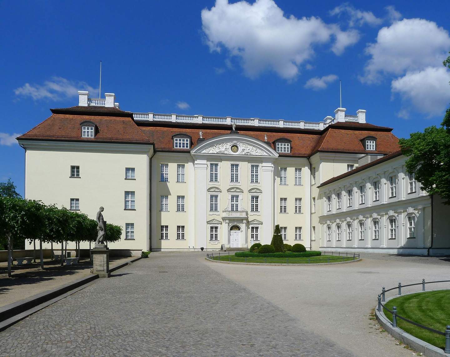 Large, elegant building with red roof and white facade, fronted by a stone path and a statue, set against a clear blue sky with few clouds.