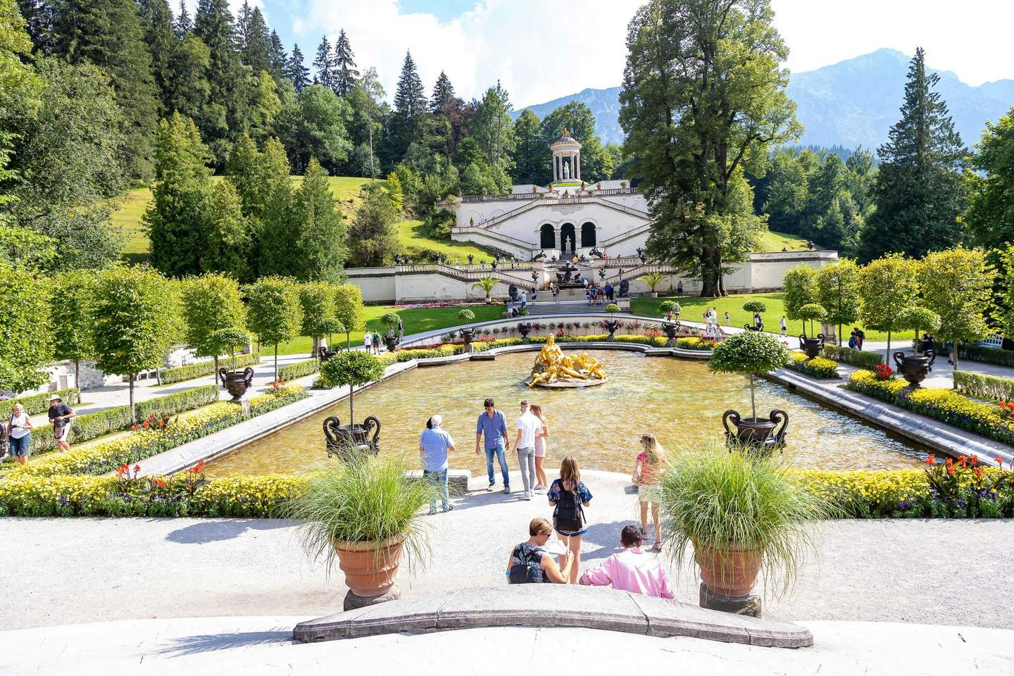 People walk near a fountain with a golden statue in a landscaped garden, leading to a white building with steps and greenery.