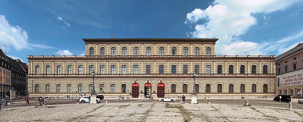 A large, historic building with arched windows and a symmetrical façade, located in an open courtyard with people and cars.