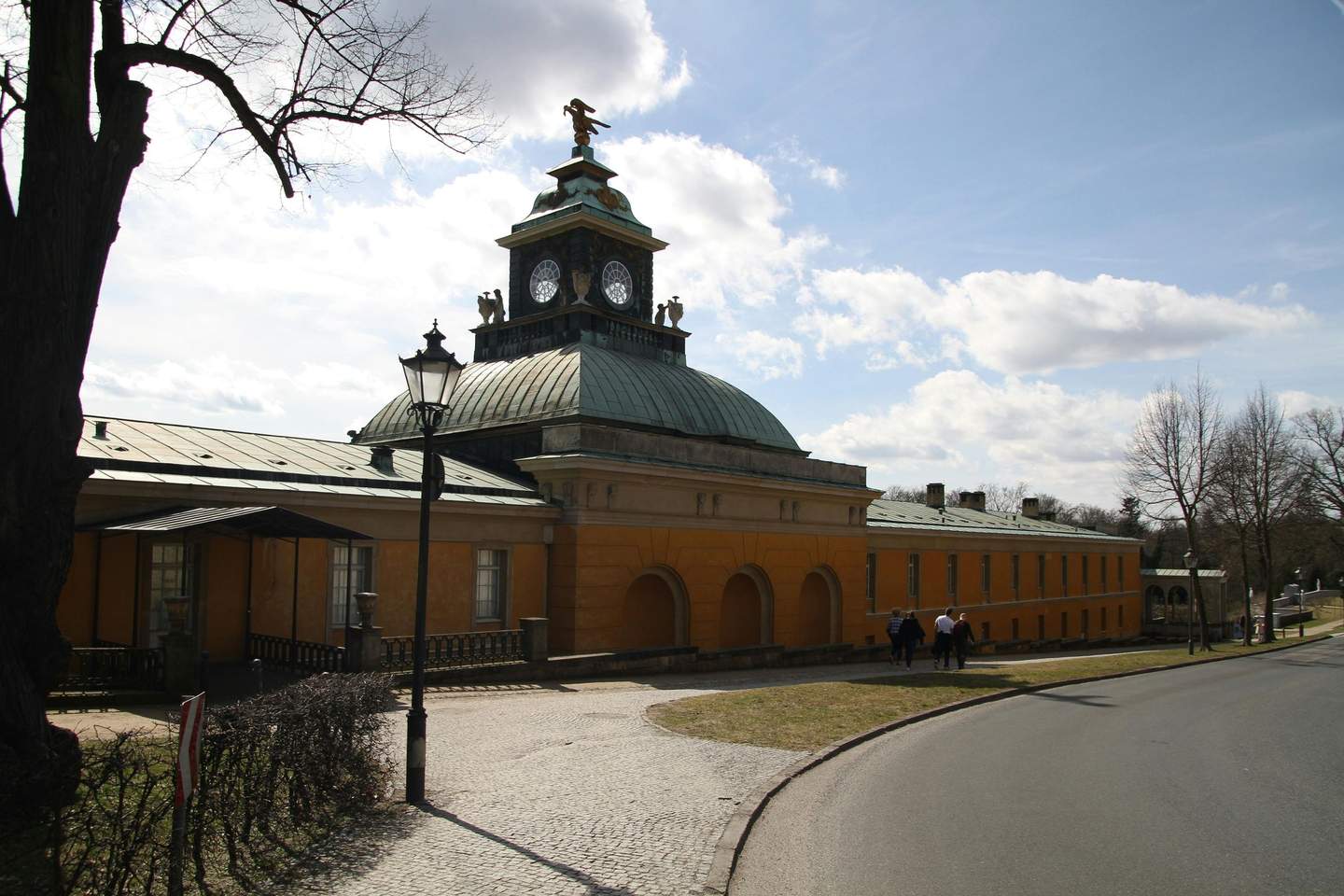 A mustard-yellow building with a clock tower and green dome, bordered by a cobblestone path and trees, under a partly cloudy sky.