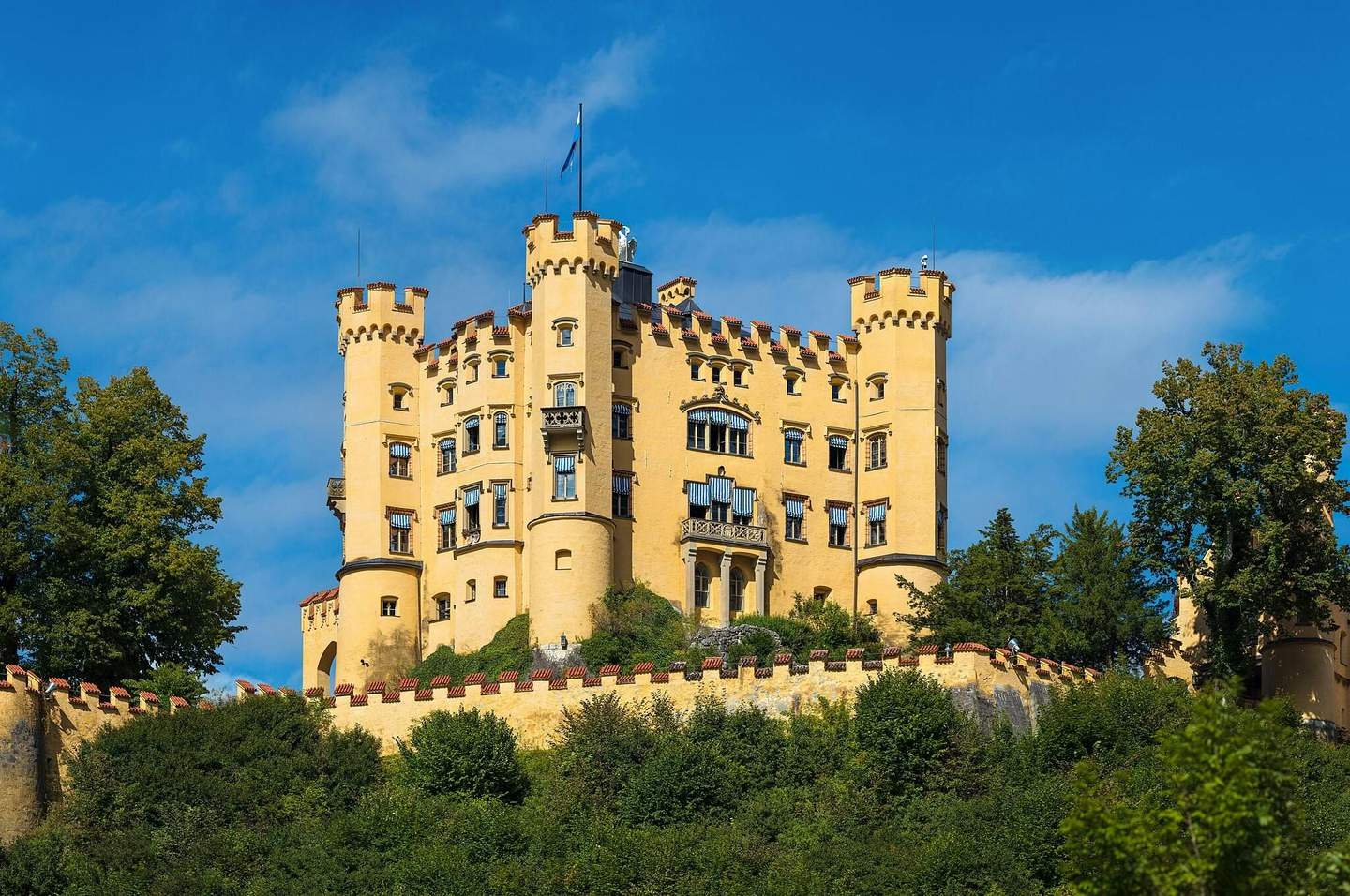 A yellow castle with multiple towers, surrounded by trees and greenery, under a clear blue sky.