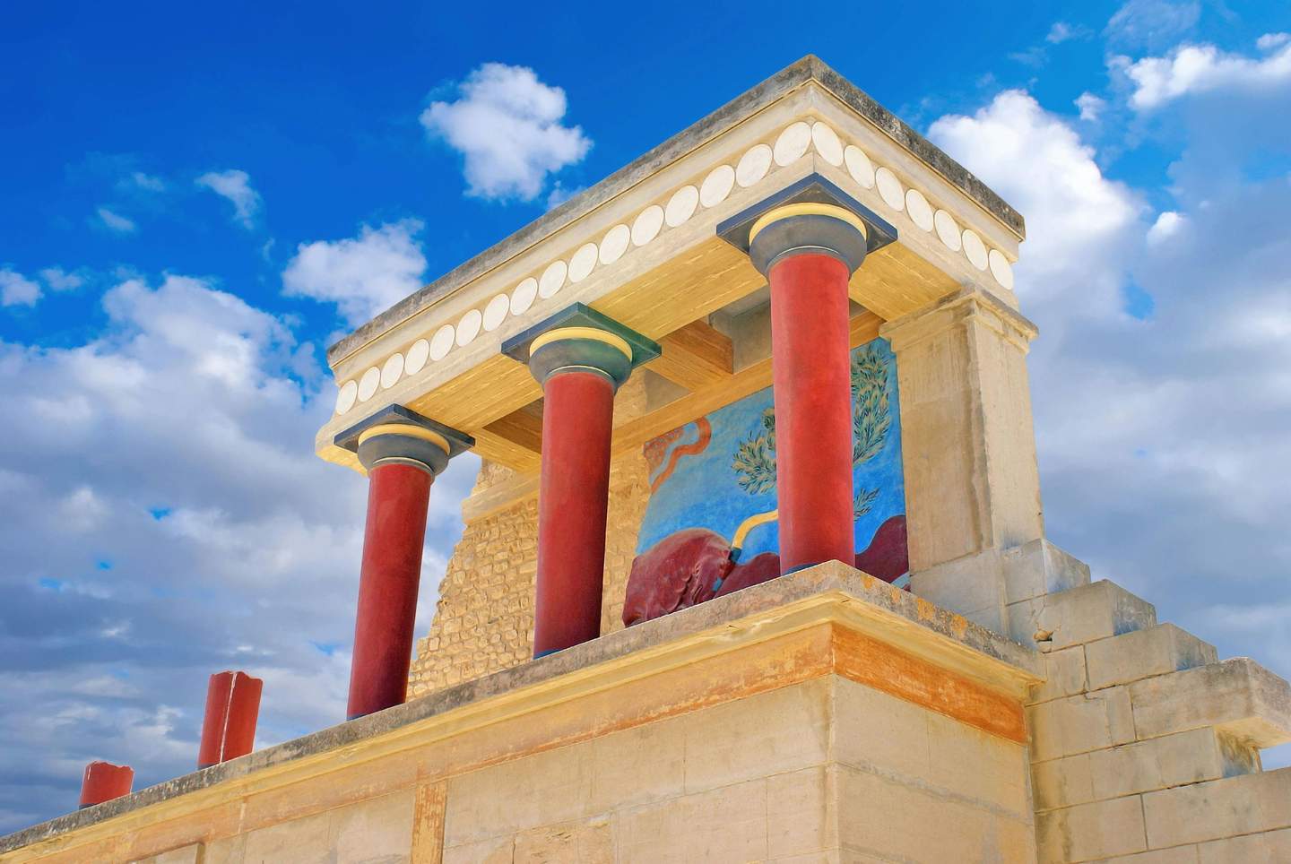 Palazzo di Cnosso Antico edificio con colonne rosse, murales dettagliati e una struttura a gradoni, sullo sfondo di un cielo blu con nuvole sparse.