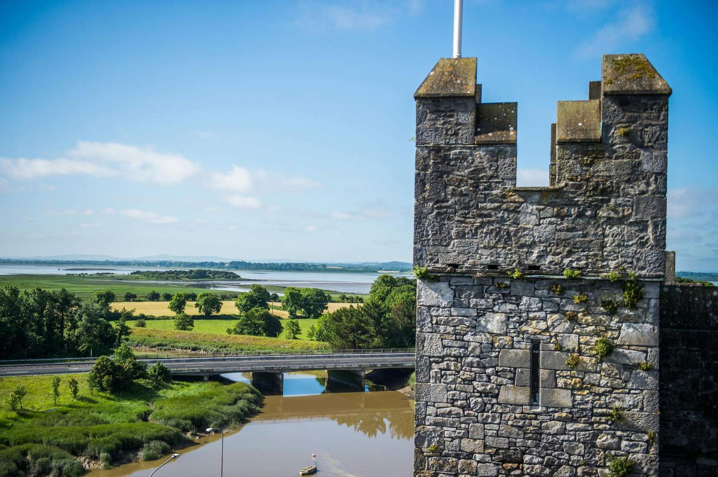 Stone tower with a crenellated top, overlooking a bridge, river, green fields, and distant trees under a blue sky.