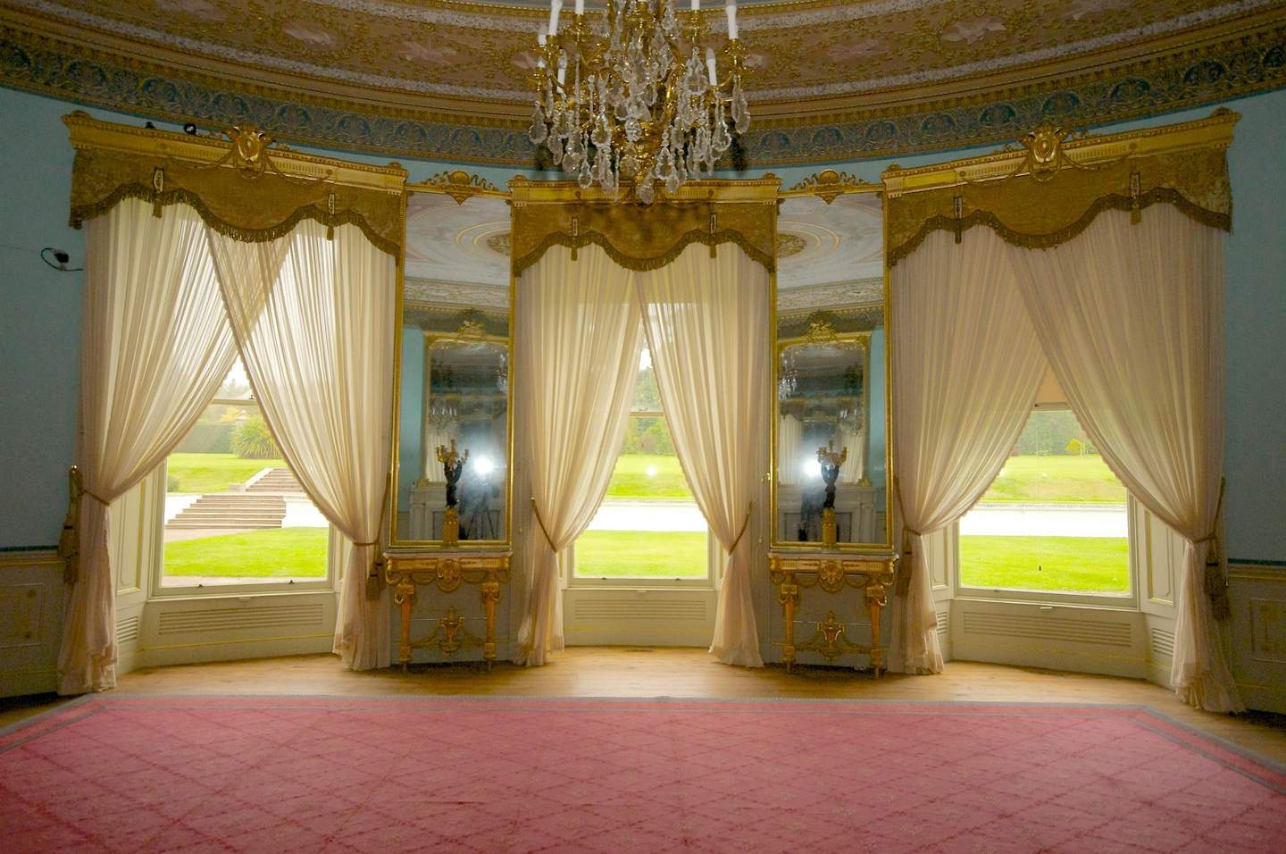 A lavish room with large windows, ornate gold trim, sheer curtains, pink carpet, and a chandelier. Green lawn visible outside.