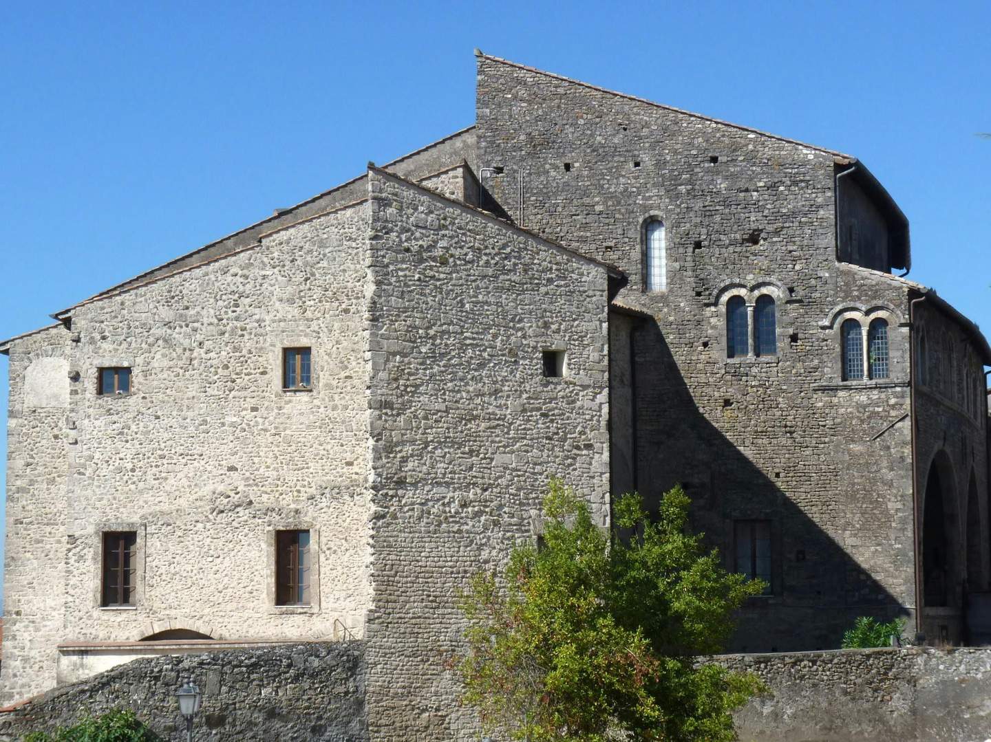 Stone building featuring arched windows, a mixture of light and dark bricks, and a small tree in front, set against a clear blue sky.