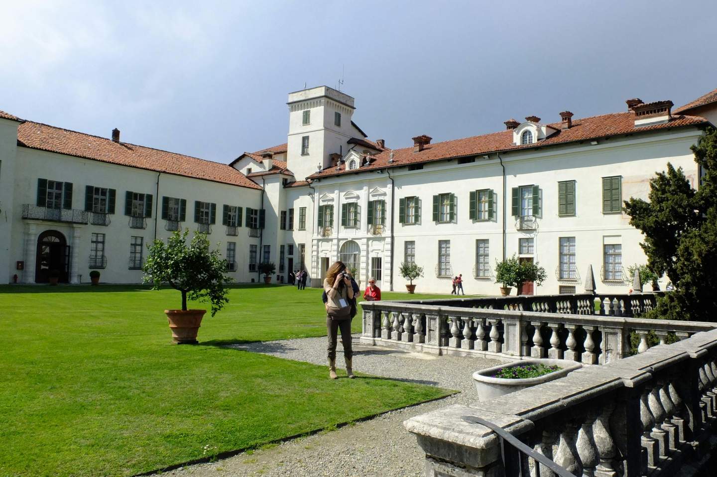 A person photographs a historic white building with green shutters, red-tiled roof, and a tower in a well-kept courtyard.