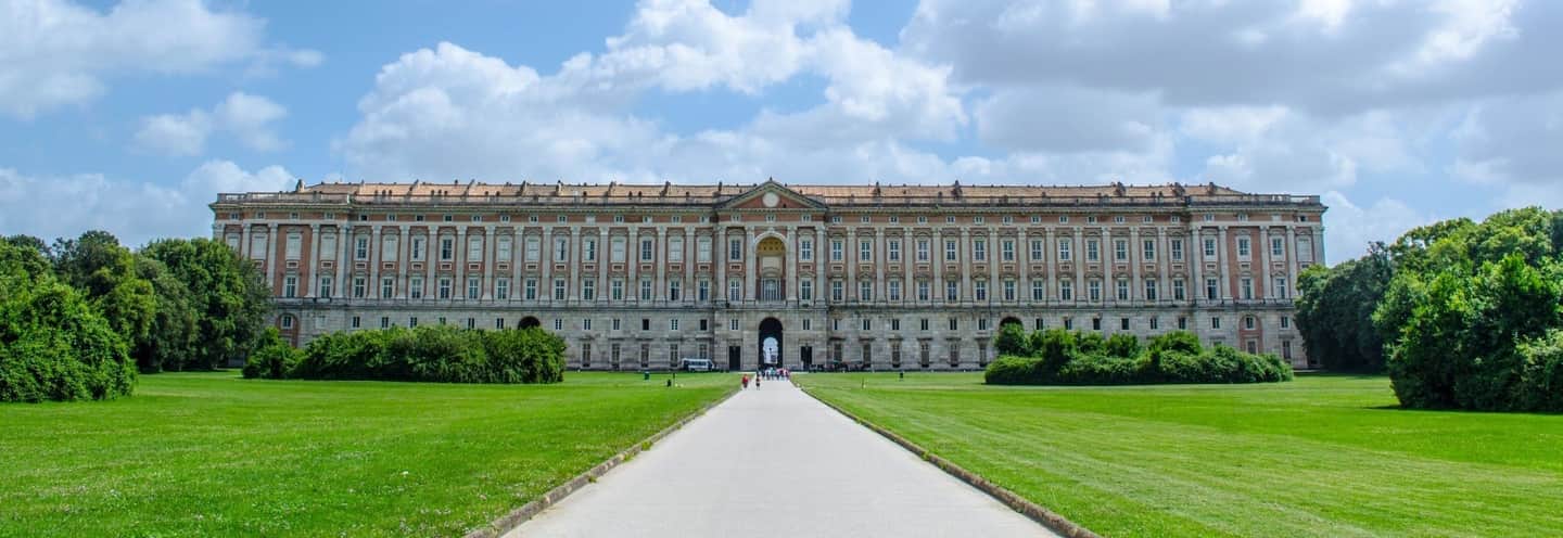 Royal Palace of Caserta An expansive palace with numerous windows and a central archway, surrounded by lush green lawns and manicured bushes.