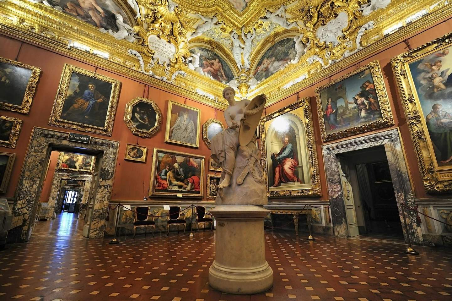 Ornate gallery with framed paintings, a large central marble statue, gold-accented ceiling, and a checkered floor.
