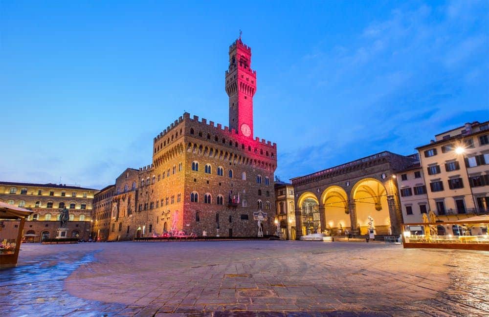 Historic stone building with clock tower and arched loggia, illuminated at twilight in an empty plaza.