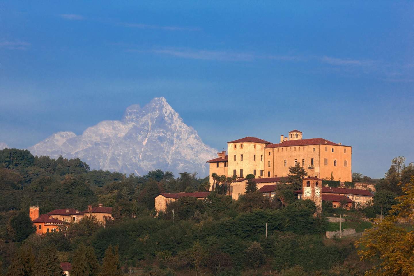 A castle with surrounding buildings on a hilltop, lush greenery, and a large mountain in the background under a clear blue sky.