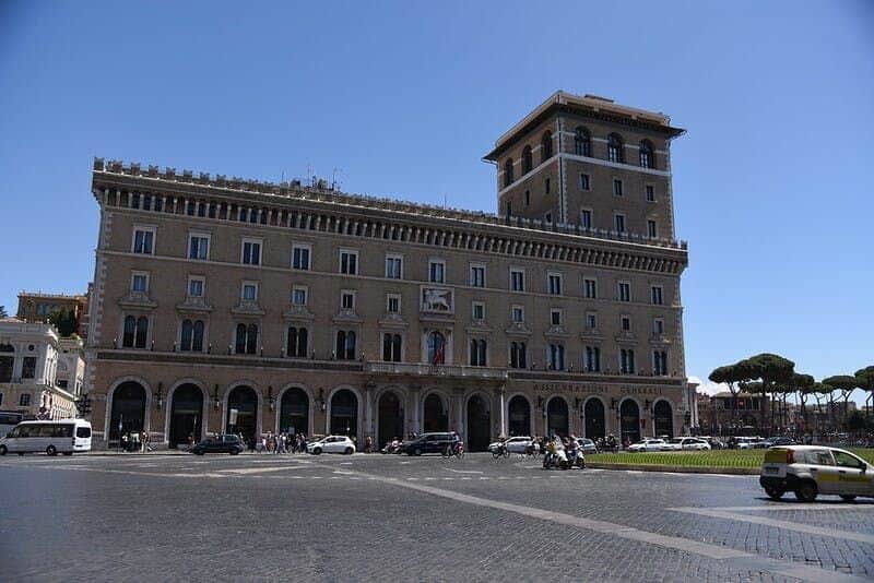 A large historic building with arches, vehicles, and people in front, under a clear blue sky. Trees and smaller buildings in the background.