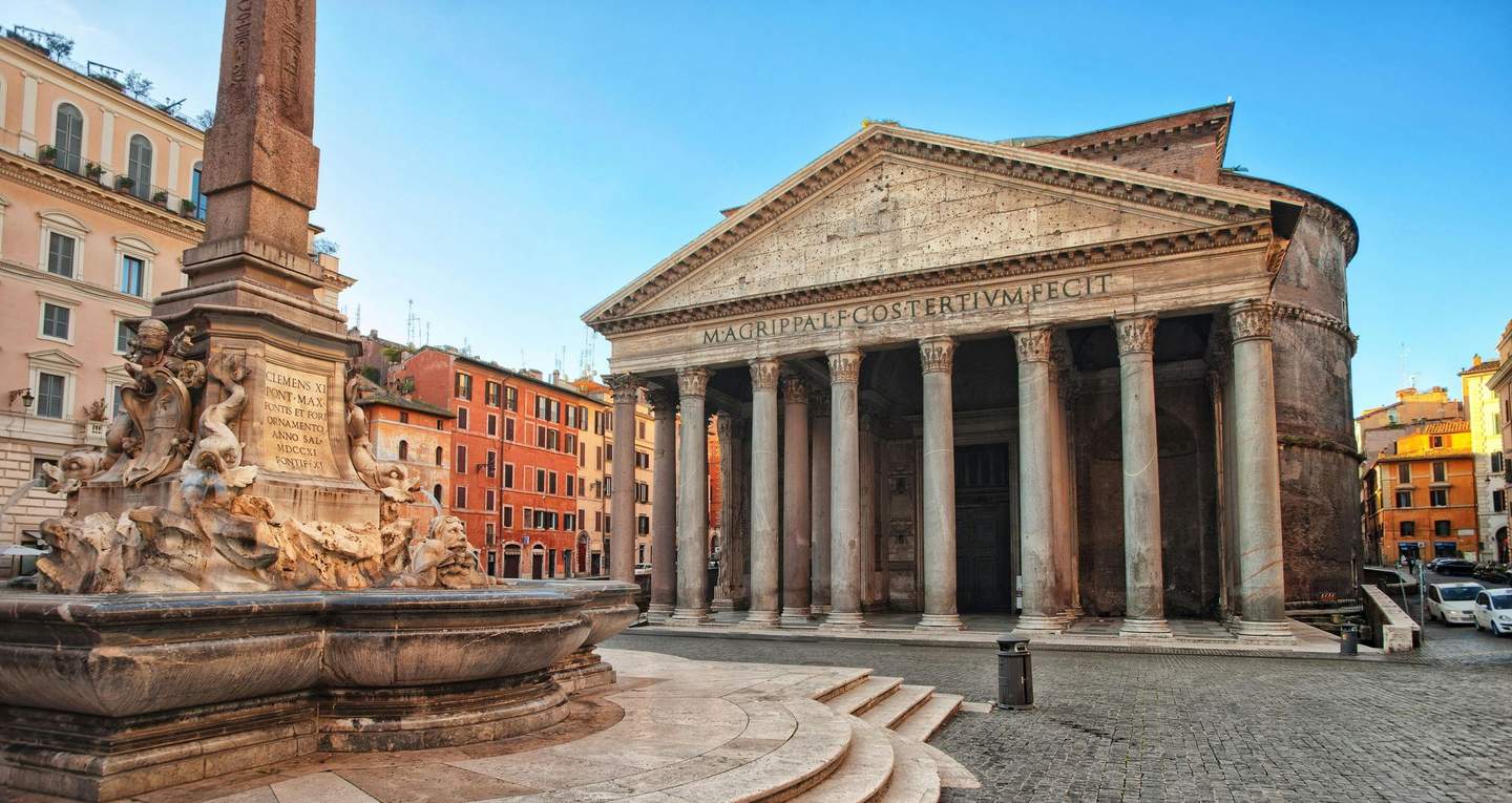 Historic stone building with large columns and a triangular pediment, adjacent to a stone fountain with sculpted figures.