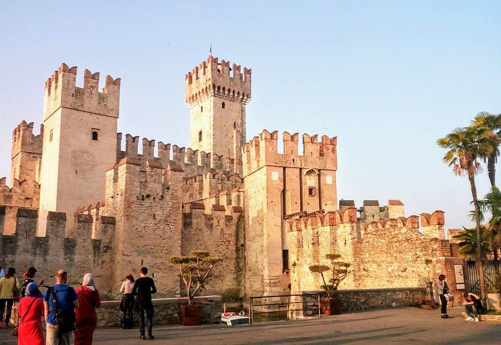Medieval castle with tall towers and crenellated walls, surrounded by people and palm trees, under a clear sky.