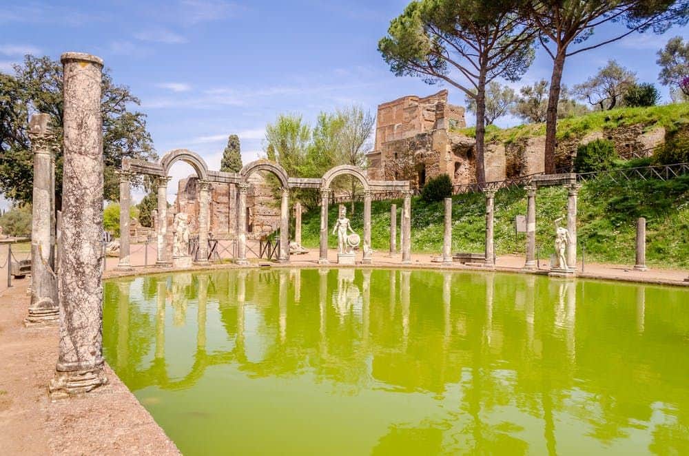 Villa Adriana Antike Ruinen mit Statuen und Steinsäulen umgeben ein grünes, spiegelndes Becken vor dem Hintergrund von Bäumen und blauem Himmel.