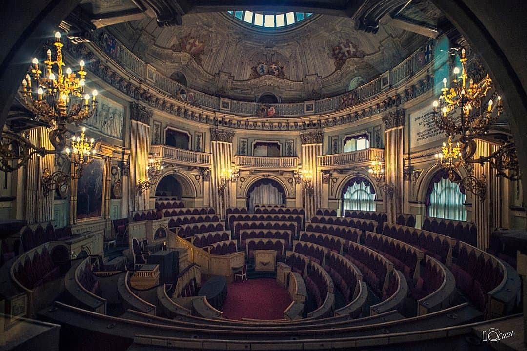 Palazzo Madama Lavish, empty legislative chamber with ornate chandeliers, red velvet seating, and a domed ceiling featuring decorative artwork.