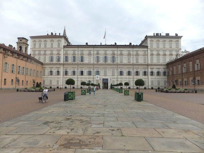 Large, ornate building with symmetrical architecture, flag on top, wide paved courtyard, people walking, and cyclists on the left.