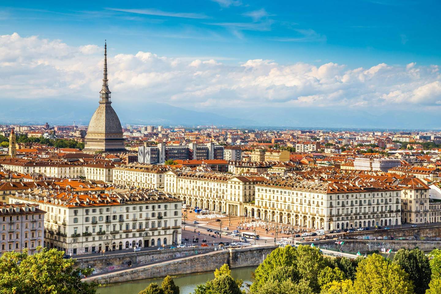 Sindone di Torino (Shroud of Turin) Cityscape with a prominent building featuring a tall spire, surrounded by historic architecture, trees, and a river in the foreground.