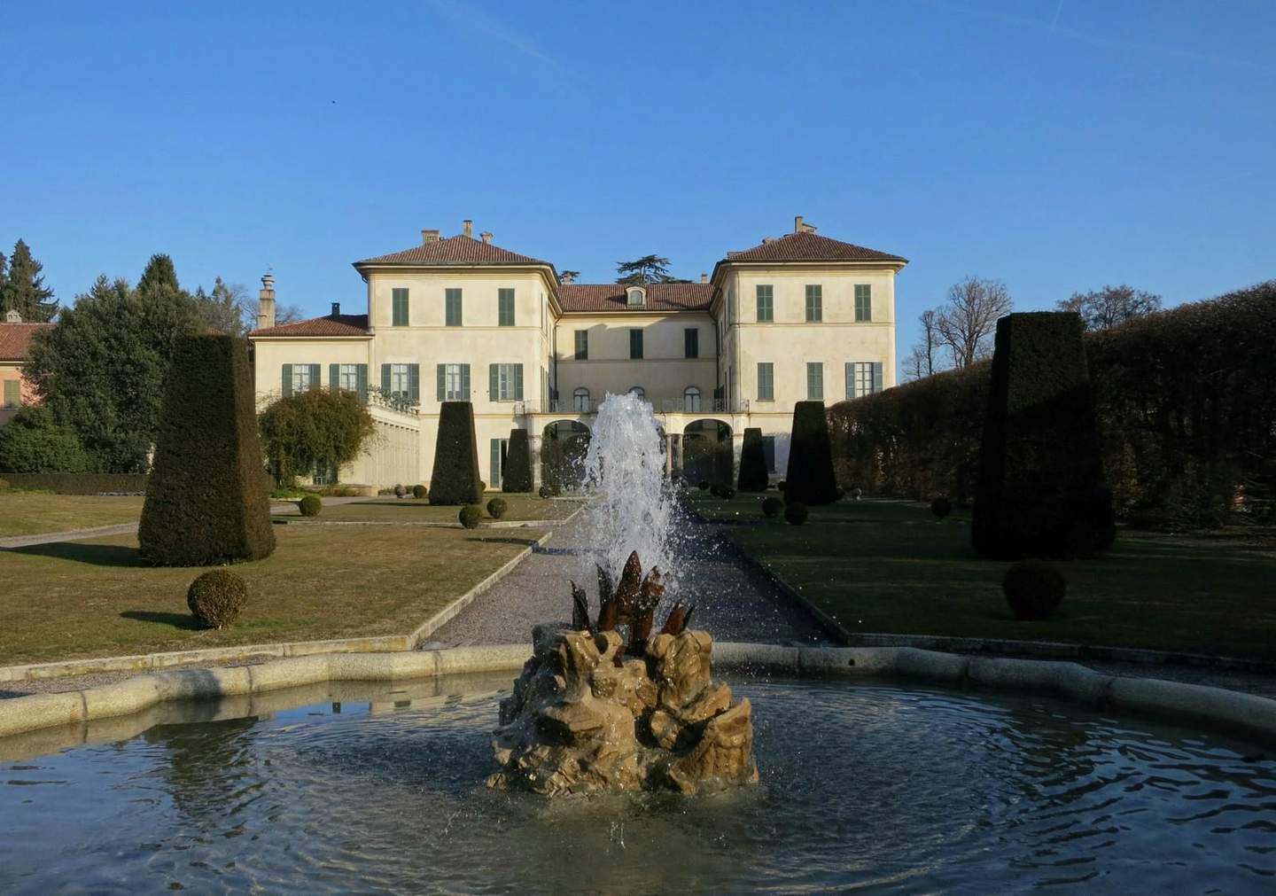 Fountain with water spraying in front of a large, cream-colored mansion with green shutters, surrounded by trimmed hedges and trees.