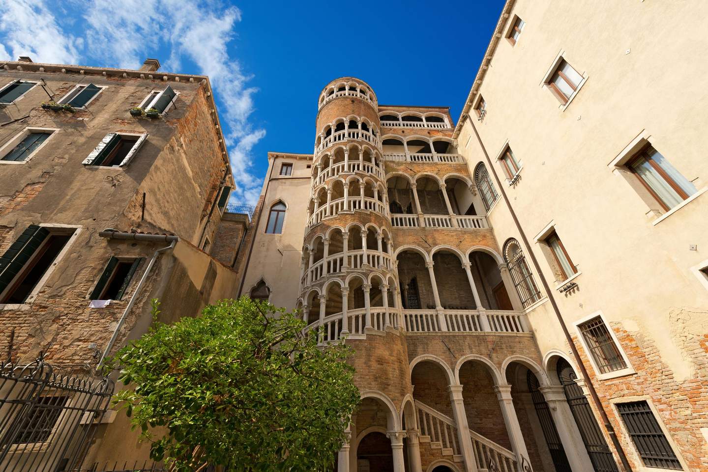 Tall Italian building with numerous arches and balconies against a blue sky, surrounded by other older buildings and a tree.