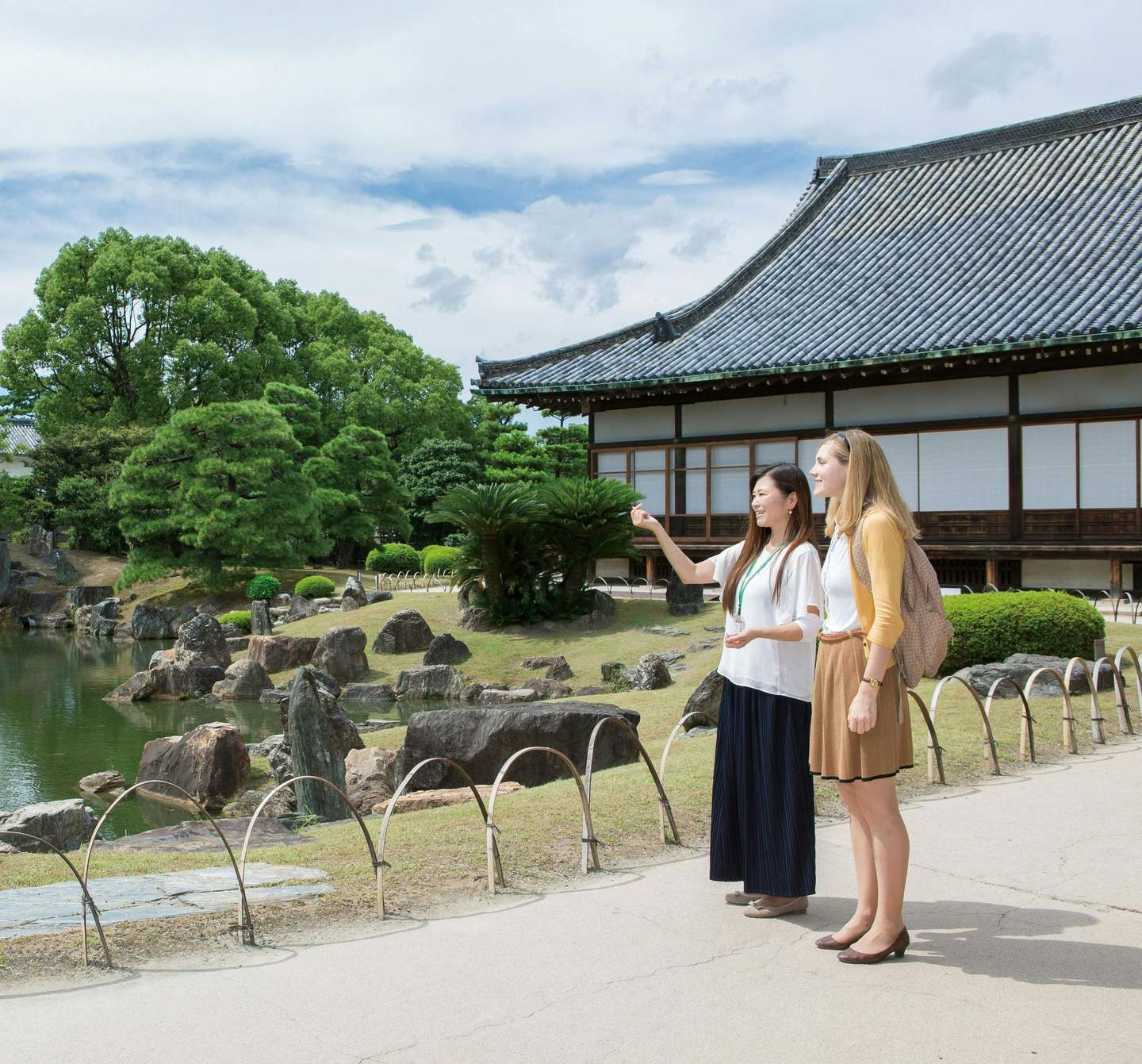 Nijo Castle Two women stand in front of a traditional Japanese building, one gesturing towards a serene garden with trees and rocks.