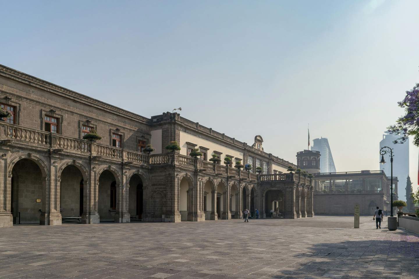 Chapultepec Castle Historic stone building with arched colonnade and potted plants, spacious courtyard, few people walking, modern skyscraper in the distance.