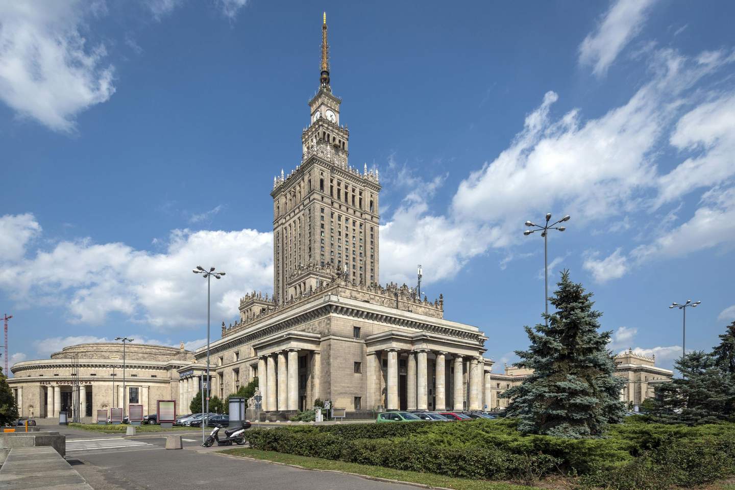 A tall, ornate building with columns and clock tower under a blue sky with scattered clouds. Trees and parked vehicles are in the foreground.