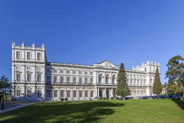 Large, white neoclassical building with multiple windows and a grand entrance, set amid a manicured lawn under a clear blue sky.