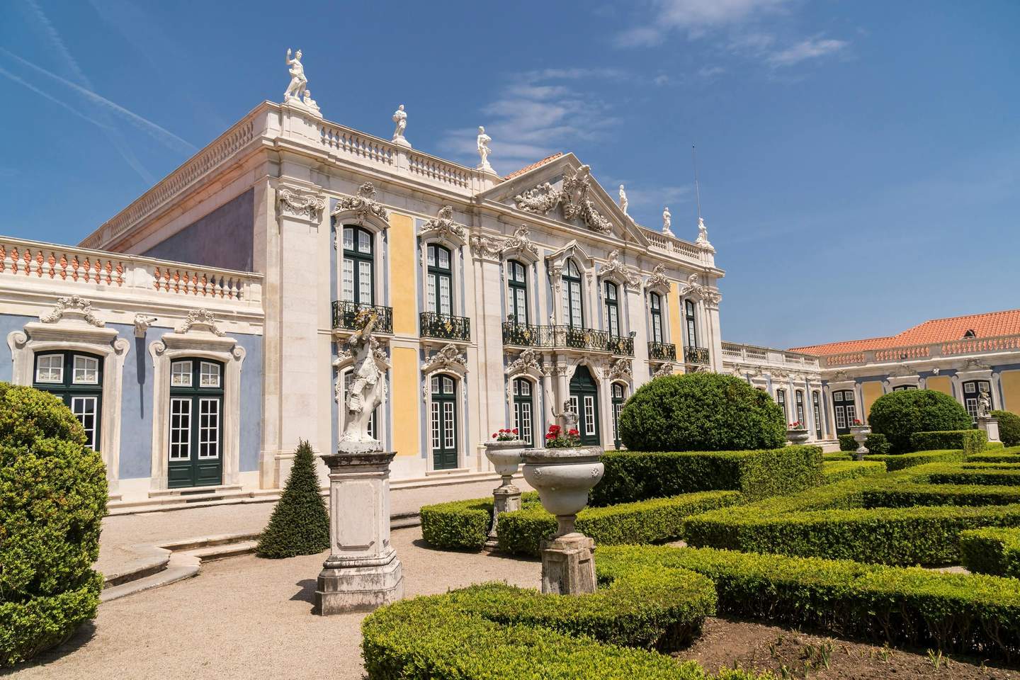 Ornate building with tall windows, detailed facades, statues, and manicured hedges in the foreground under a clear blue sky.