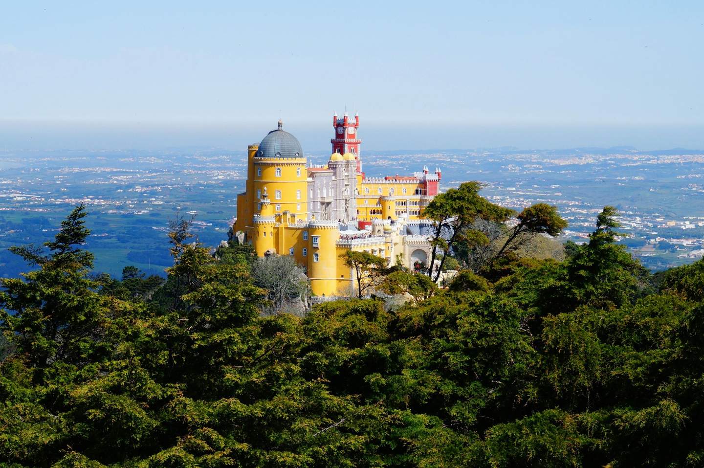 National Palace of Pena & Park A colorful, ornate castle with yellow, red, and white towers sits atop a lush, green hill with a vast landscape in the background.