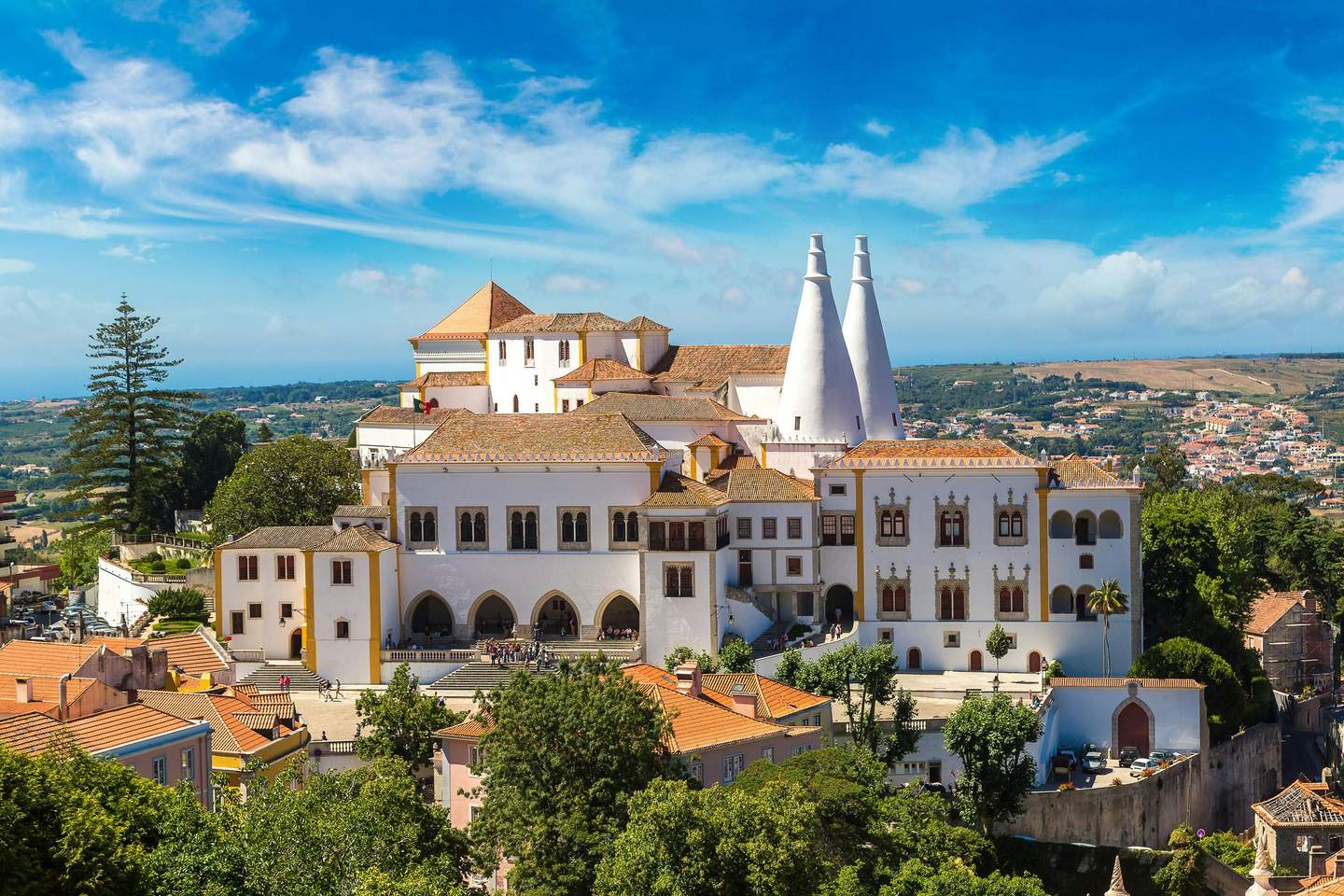 Hilltop palace with two conical chimneys, surrounded by lush greenery and neighboring buildings, under a bright blue sky.