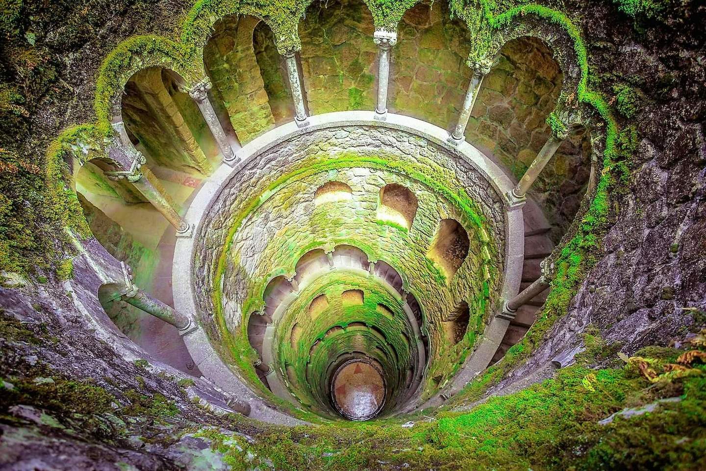 A moss-covered, spiral stone staircase descends into a circular well, with arches and columns lining the walls.