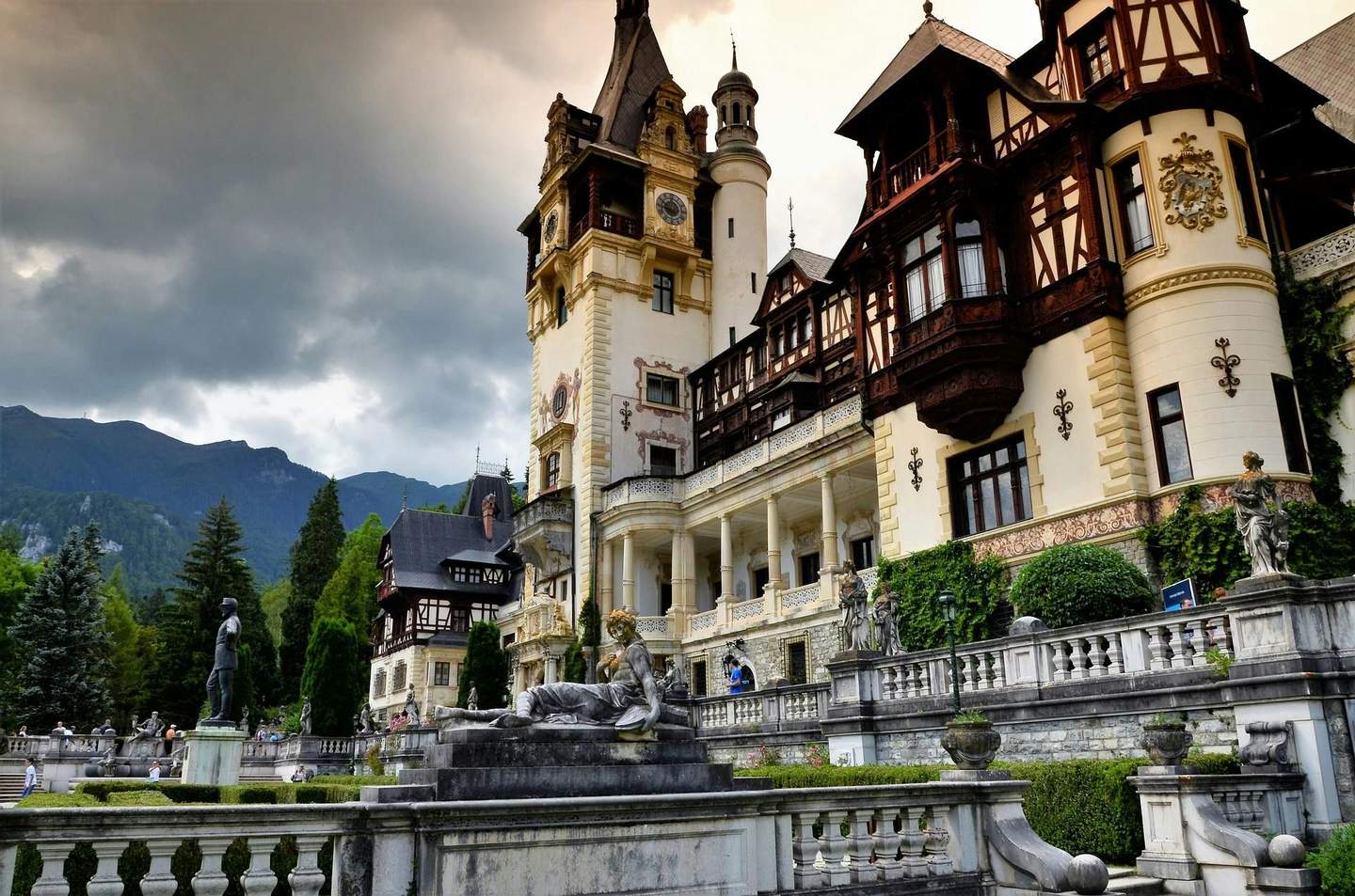 A grand palace with ornate towers and sculptures, set against a backdrop of cloudy sky and forested mountains.