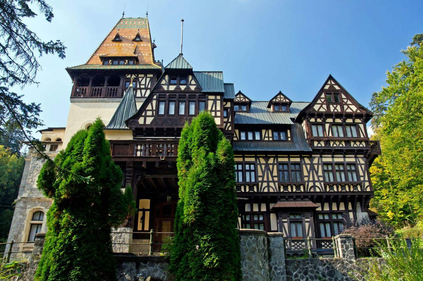 Large, ornate Tudoresque building with intricate wood detailing and tall, peaked roof surrounded by trees under a clear blue sky.